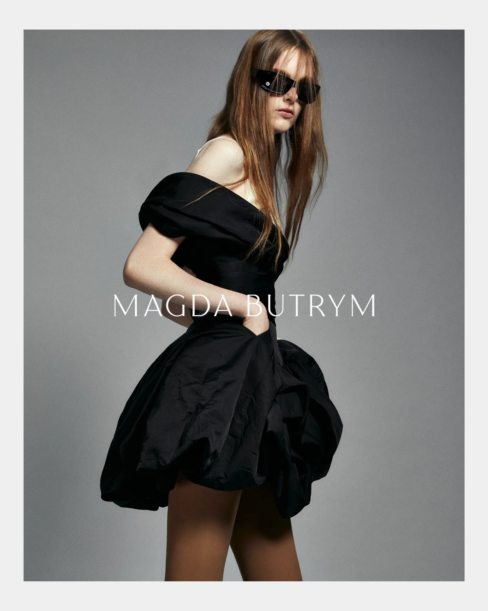 Model wearing a short black off-shoulder dress and sunglasses against a gray background.