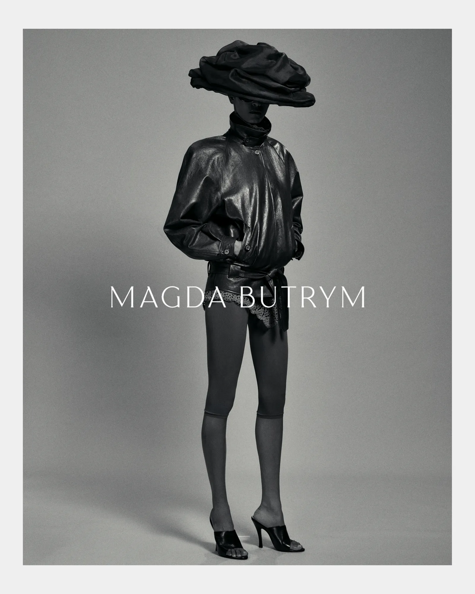 Model wearing oversized black leather jacket, lace shorts, sheer tights, and large sculptural hat.