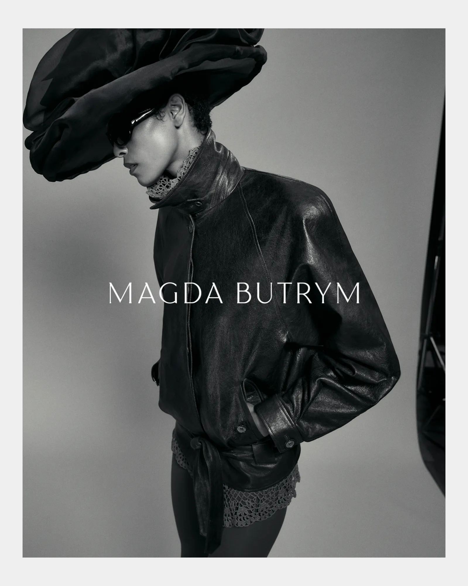 Profile of model in oversized leather jacket, large hat, lace collar, and sunglasses on gray backdrop.