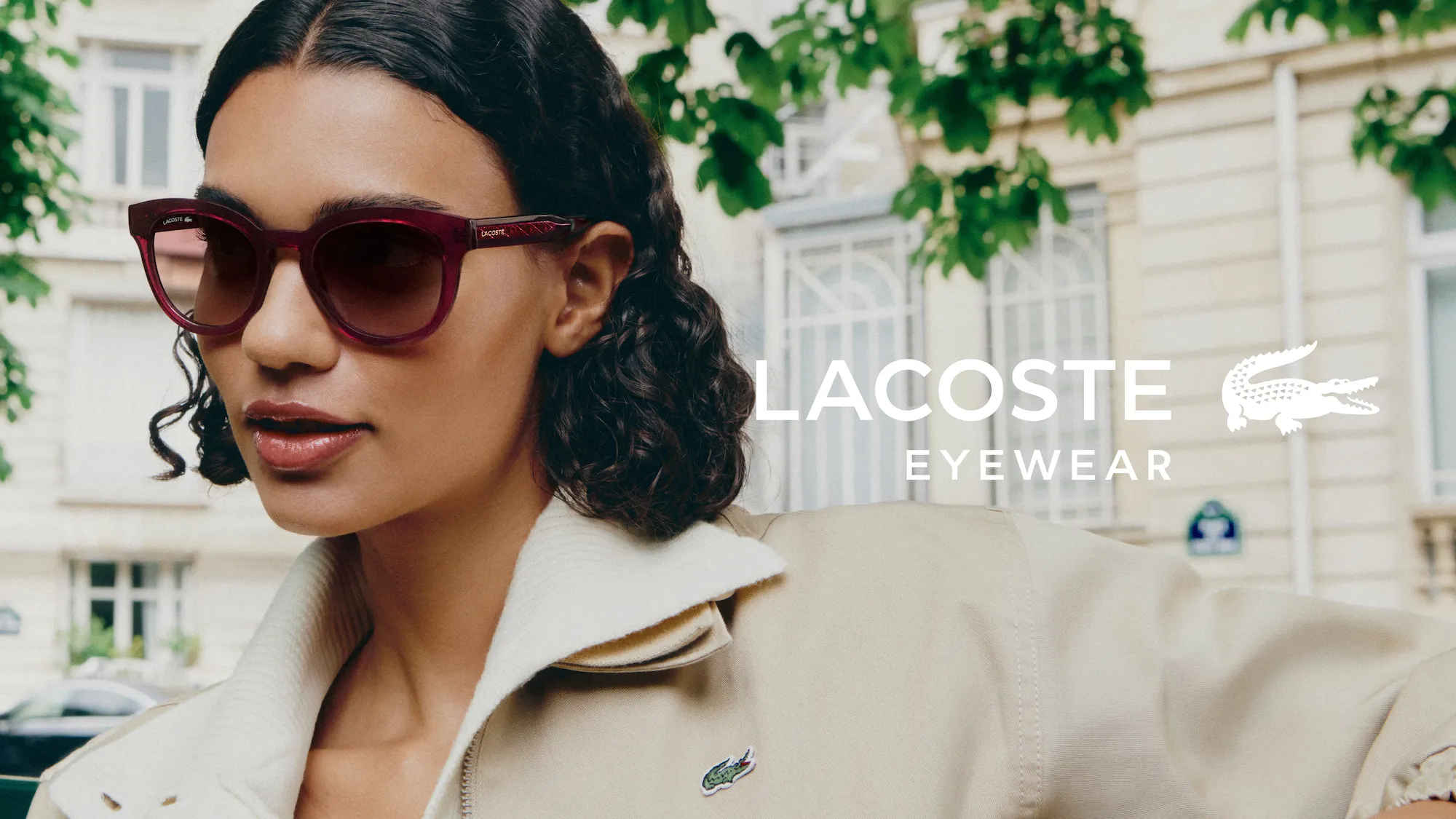 Woman wearing burgundy Lacoste sunglasses and a beige jacket with Lacoste logo outside.
