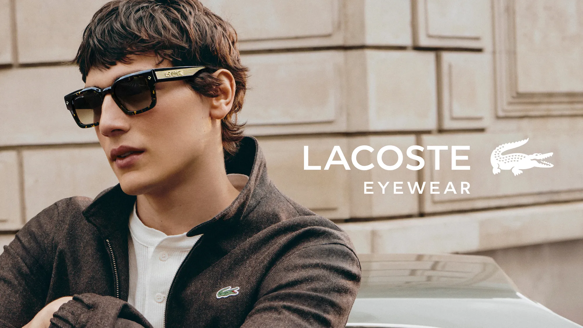 Young man wearing dark Lacoste sunglasses and jacket posing against beige stone wall.