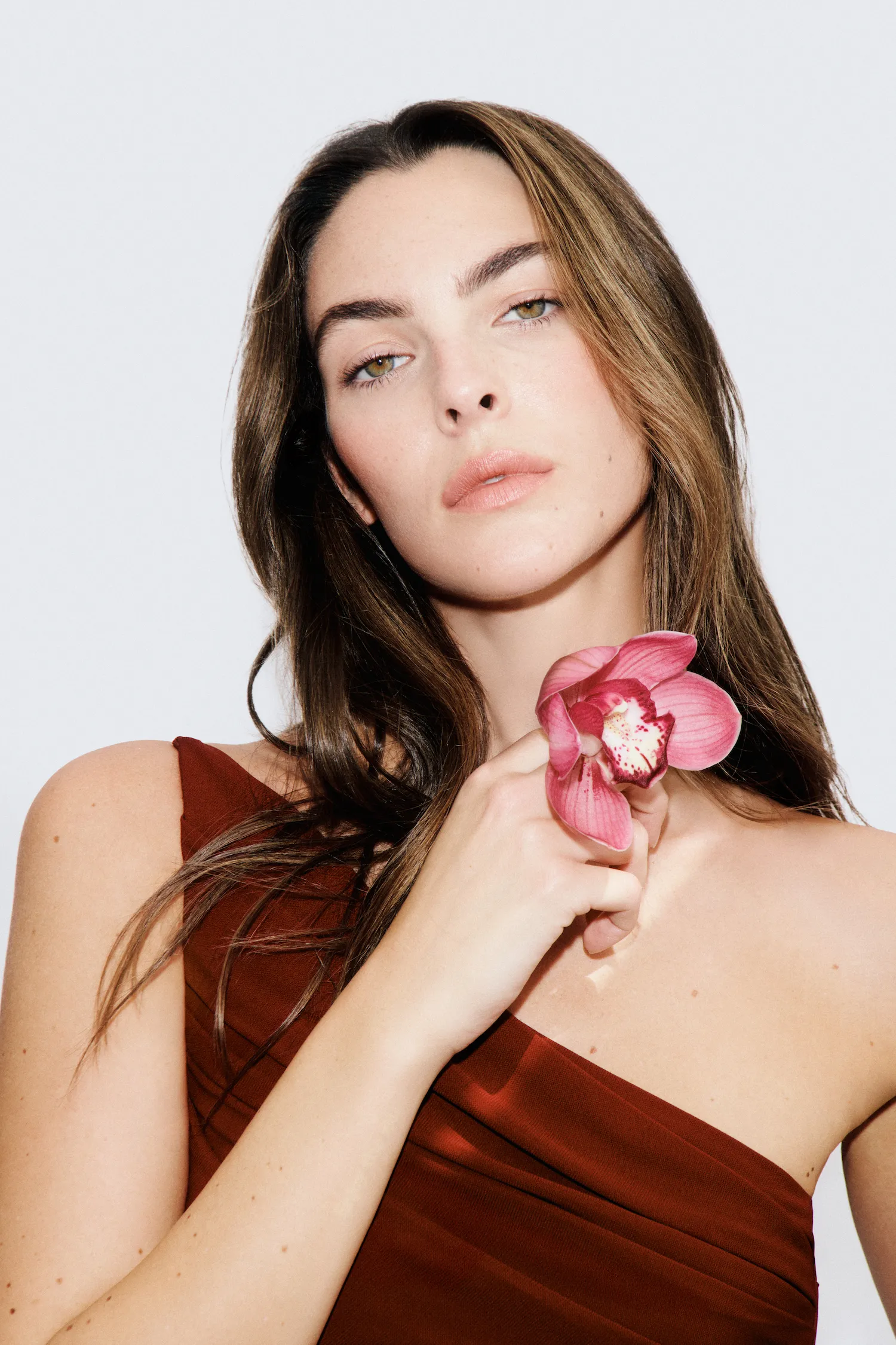 Woman in a brown one-shoulder dress holding a pink flower near her chin against a white background.
