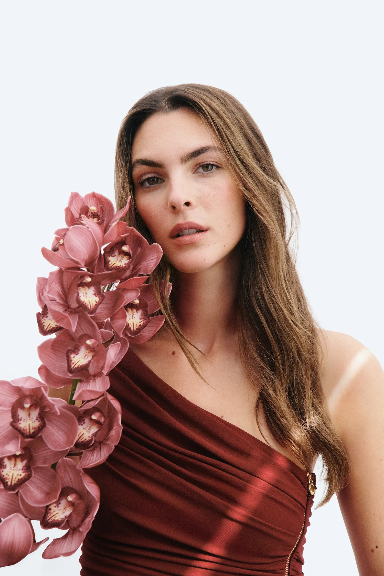 Portrait of a woman in a maroon dress holding a bunch of pink orchids against a white background.