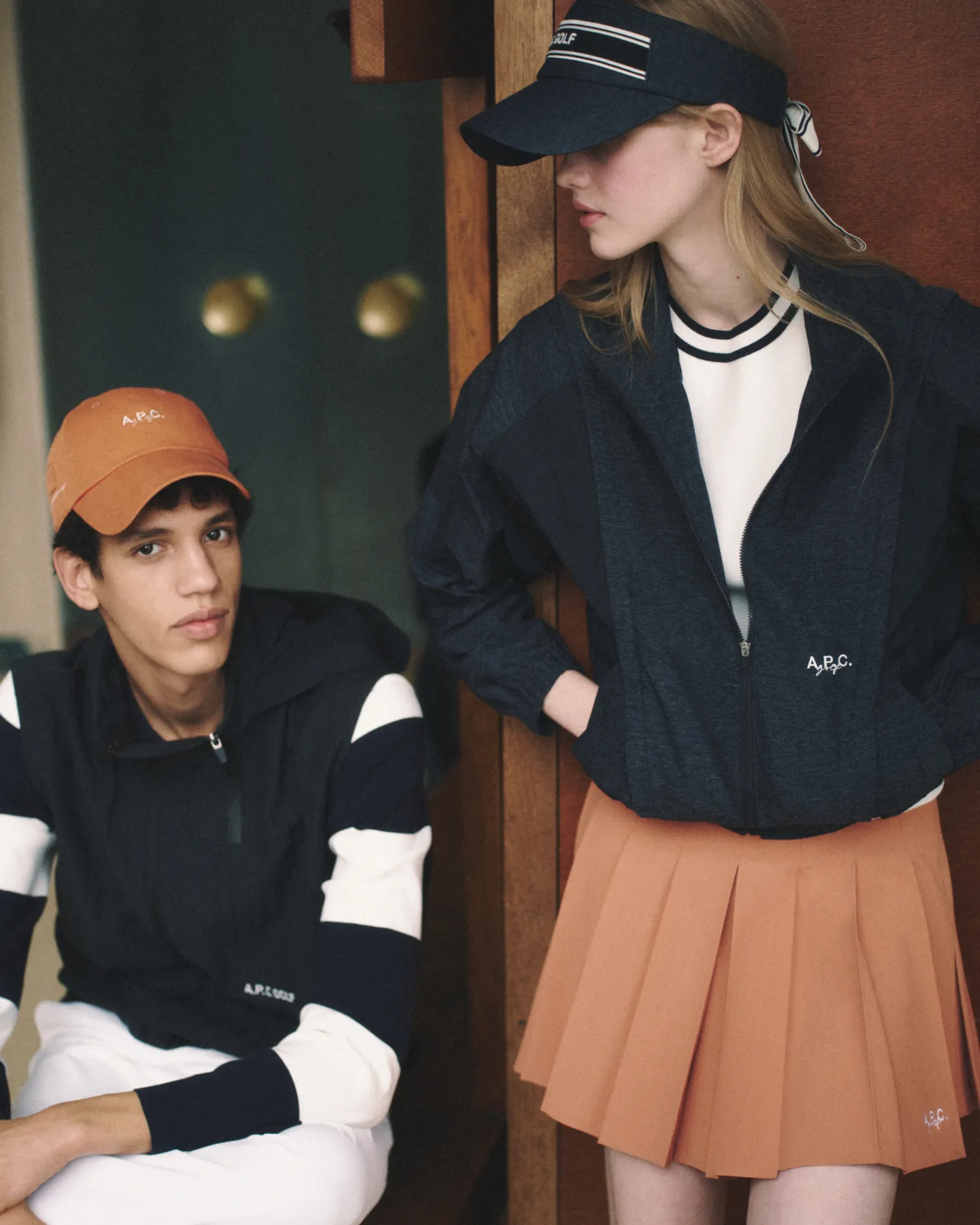 Young man seated wearing black and white outfit with orange cap, woman standing in navy jacket and orange skirt.
