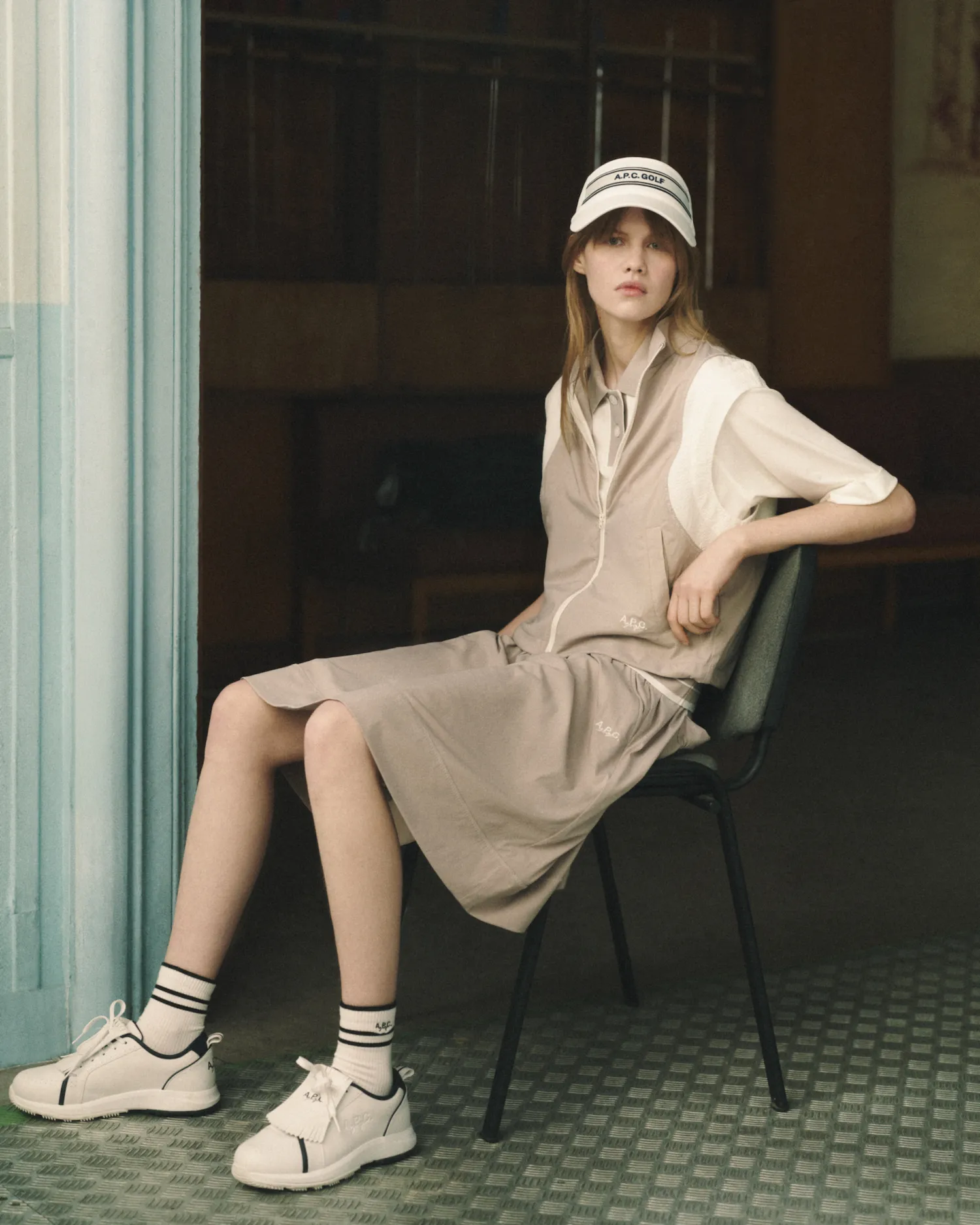 Woman sitting on chair wearing beige vest, skirt, white shirt, A.P.C. Golf cap, and white sneakers with socks.