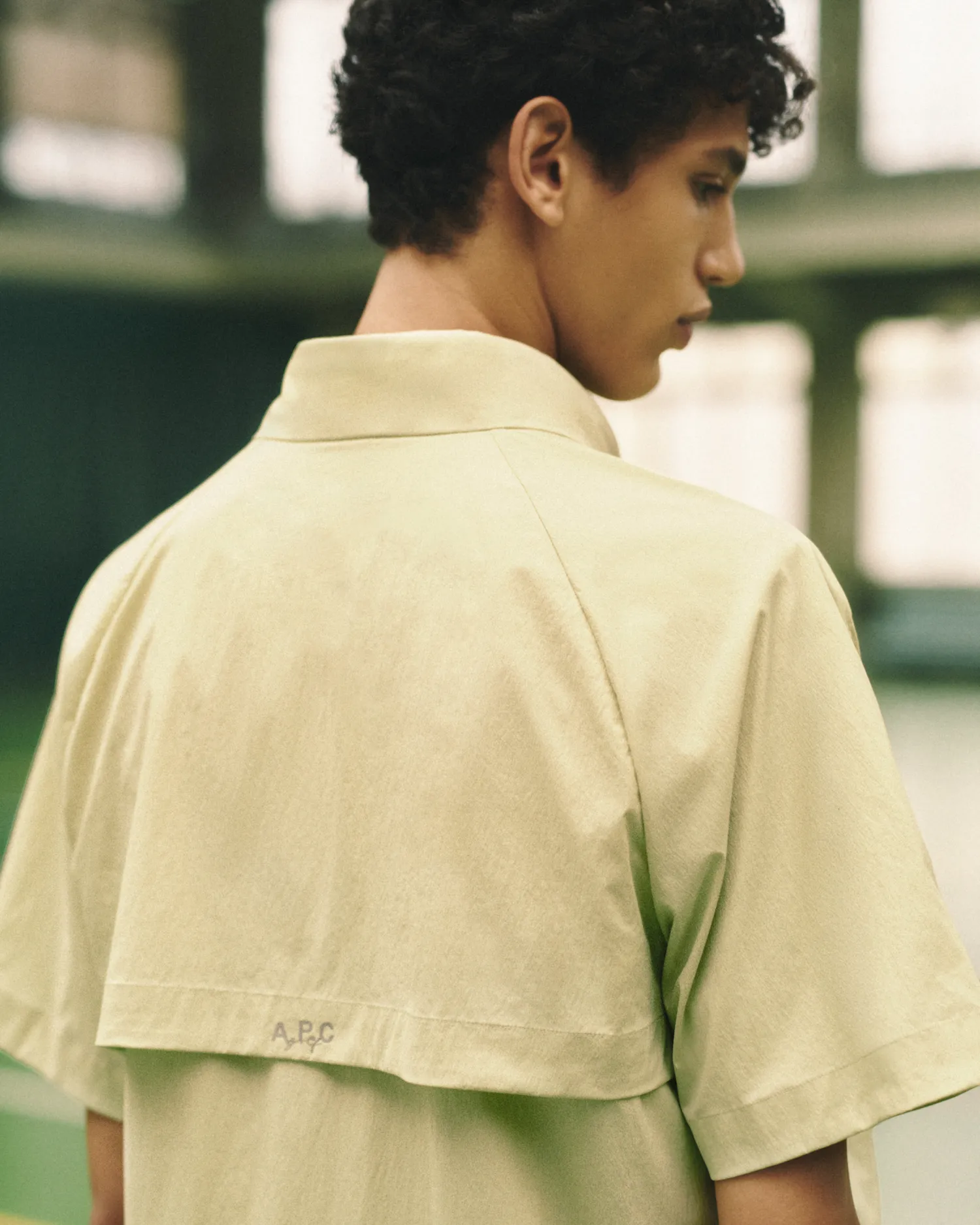 Close-up of a person in a beige short-sleeve shirt with a collar flap and A.P.C. logo on the back.