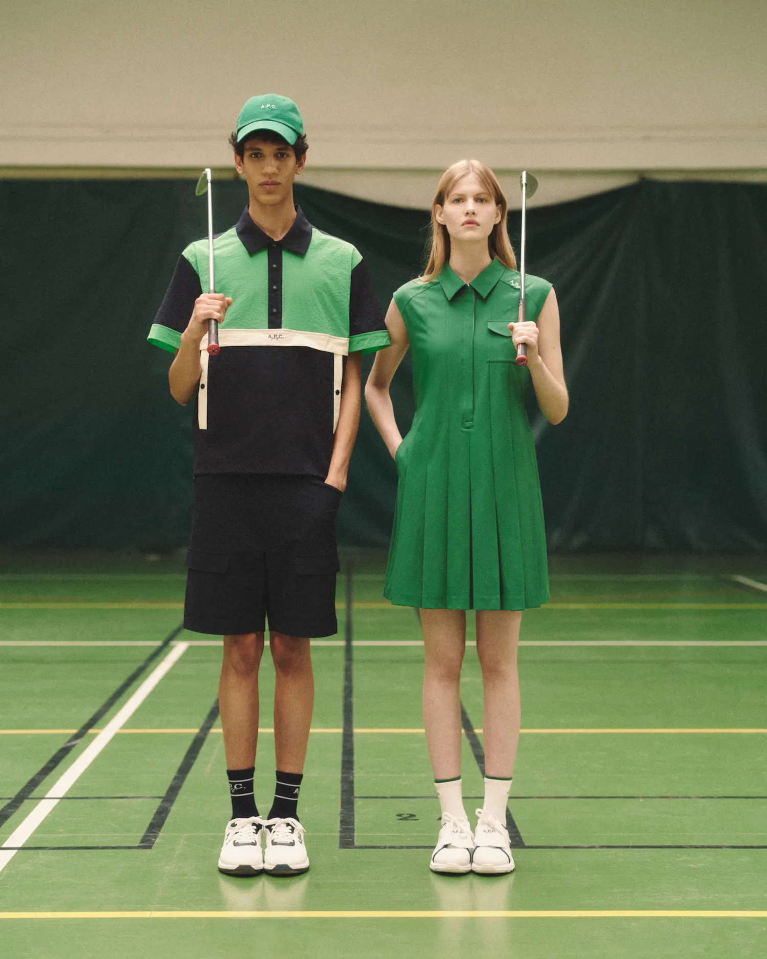 Man and woman standing side by side in green sportswear holding golf putters on indoor court.