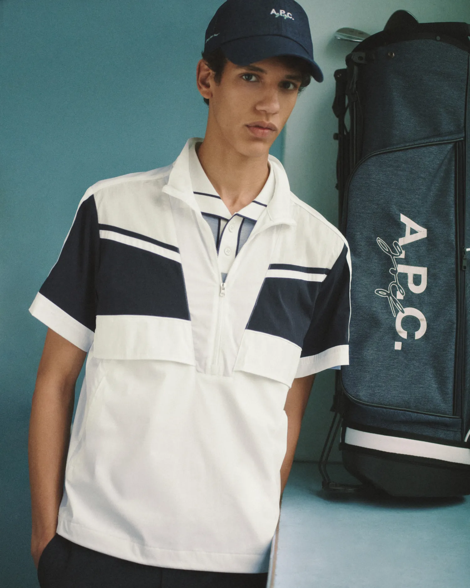 Young man wearing a black and white zip-up shirt and a navy A.P.C. cap standing next to a golf bag.