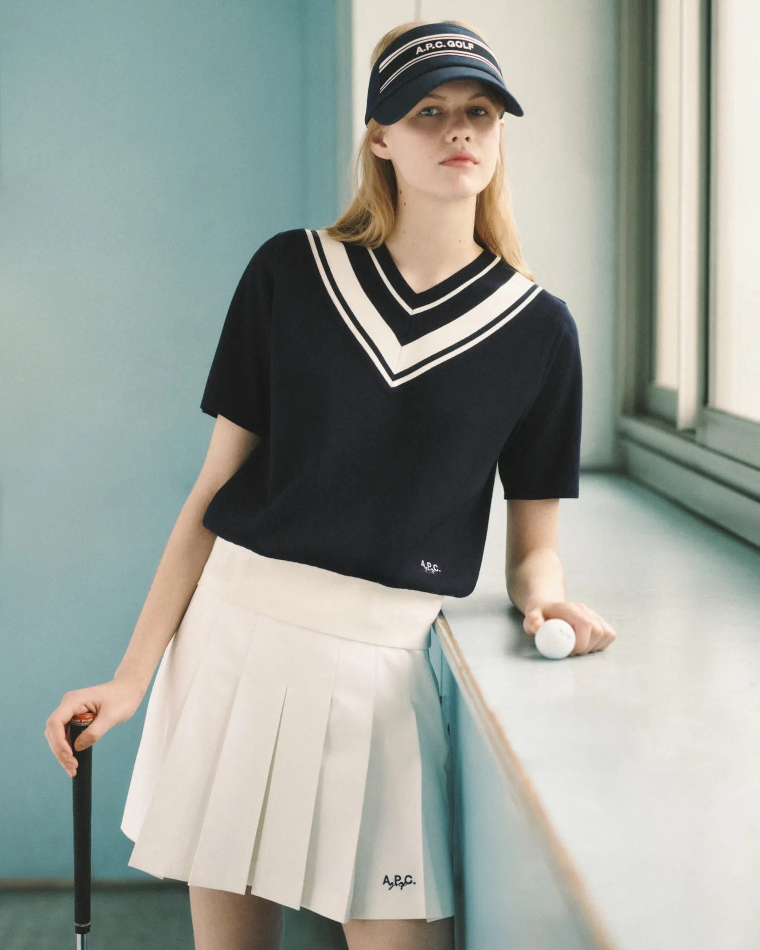 Young woman in navy golf shirt and white pleated skirt holding golf club and ball by window.