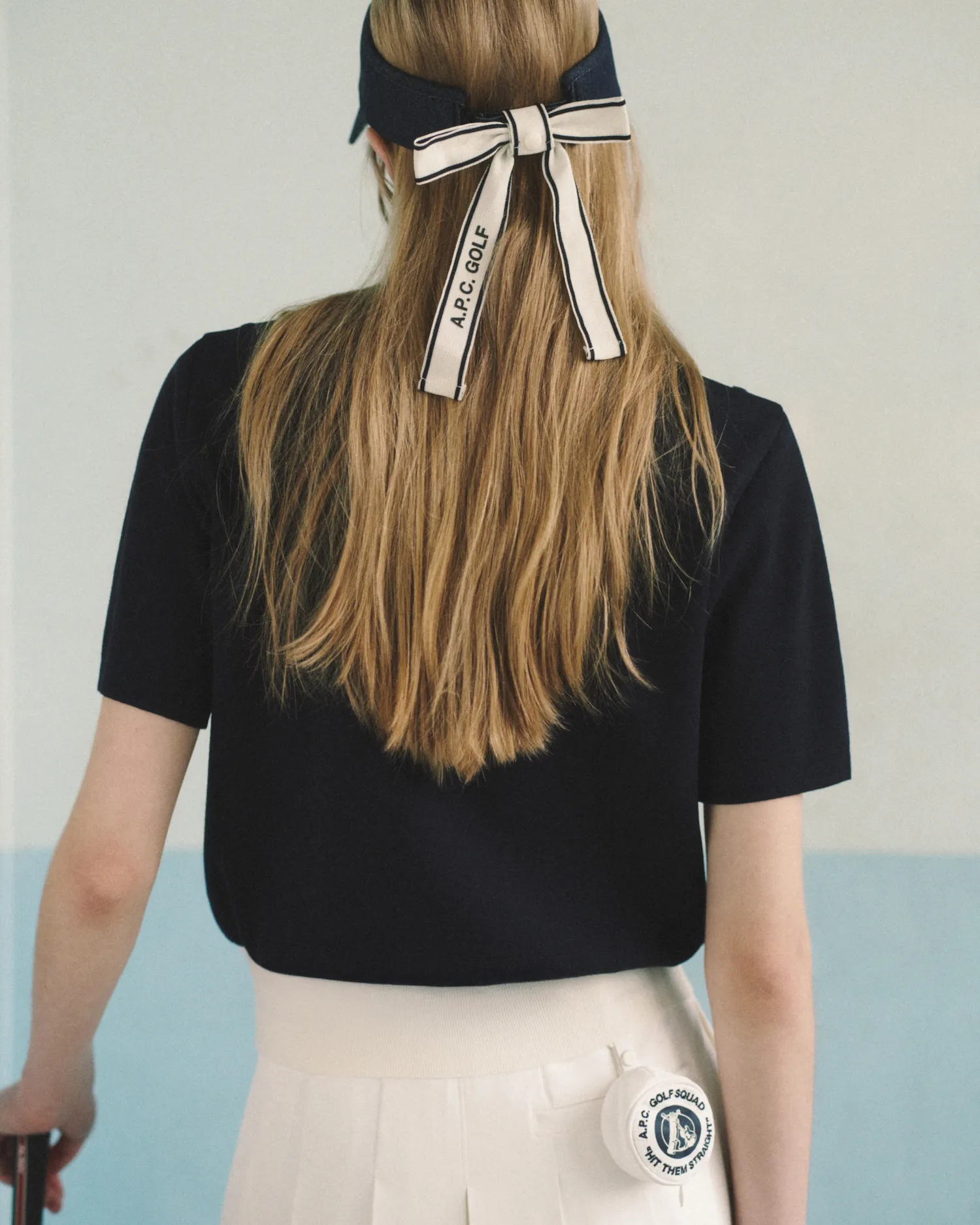 Person seen from behind wearing a black shirt and white skirt with a golf visor and ribbon tied in hair.