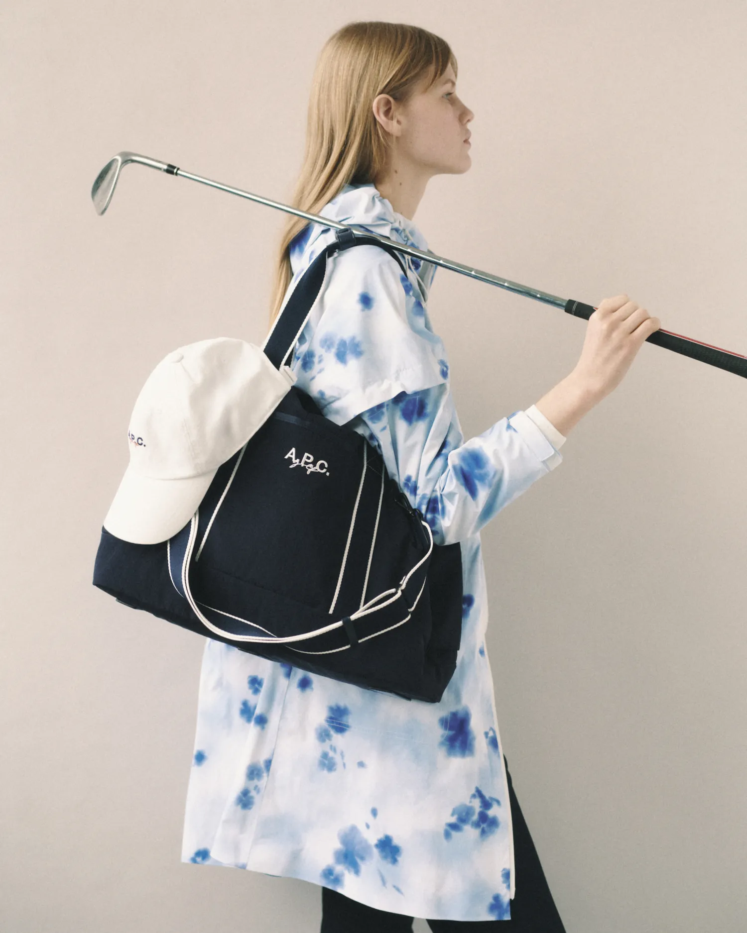 Person in white and blue tie-dye jacket carries navy bag with cap and golf club over shoulder.