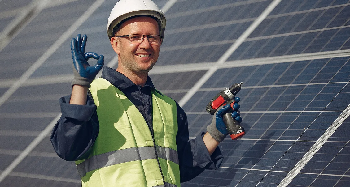 Smiling worker in safety vest and helmet holding a power drill and making an OK gesture in front of solar panels.
