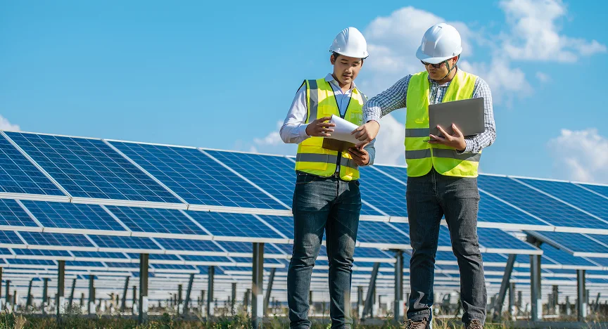 Two engineers in yellow safety vests and white helmets inspecting documents and a laptop at a solar panel farm under a blue sky.