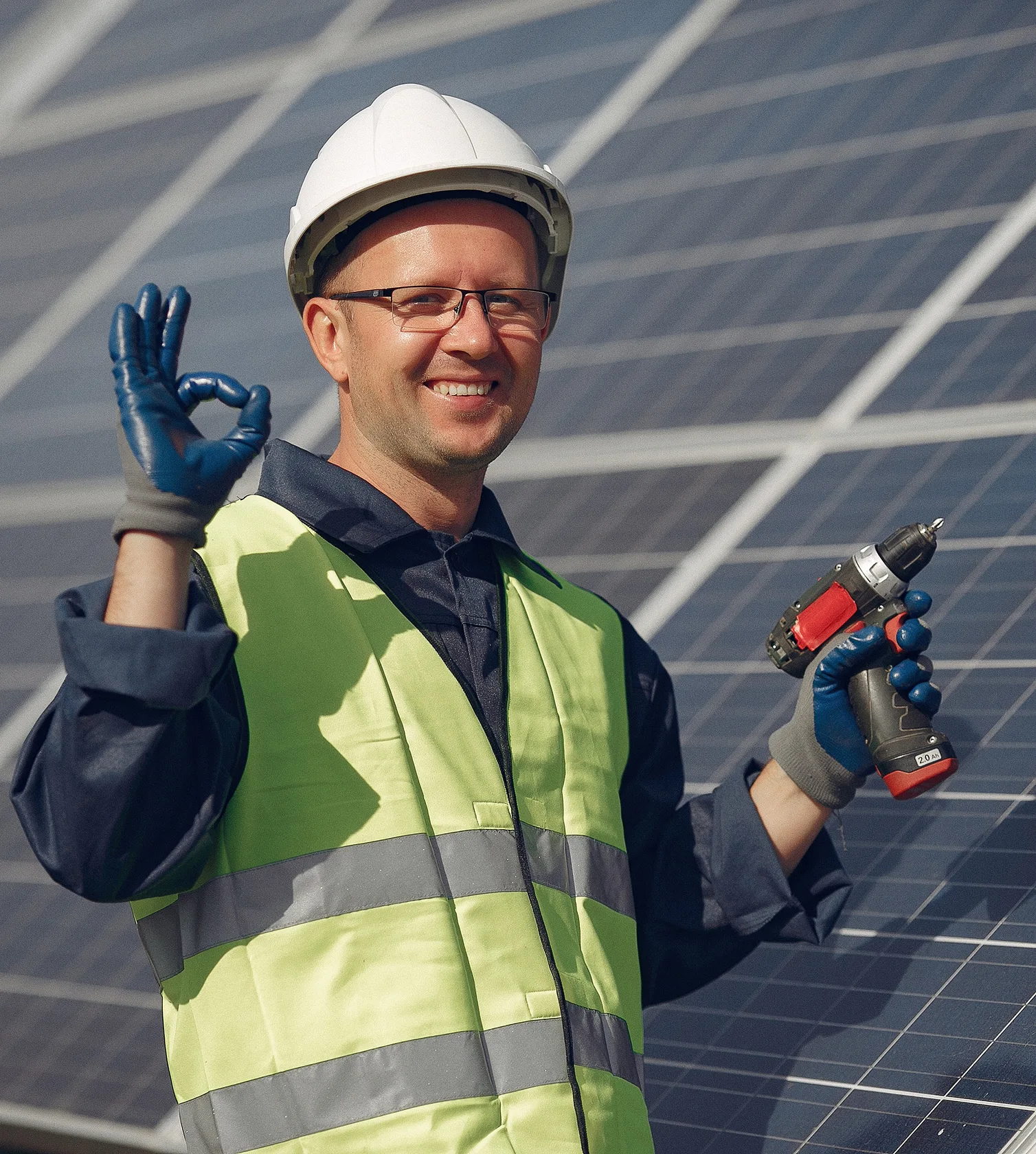 Smiling male worker in a white hard hat and yellow safety vest holding a cordless drill and making an OK hand sign in front of solar panels.
