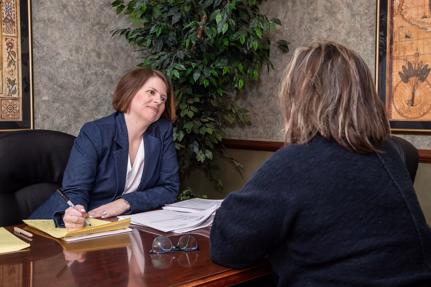 Woman in blazer writing on yellow legal pad while talking with another woman across desk in office.