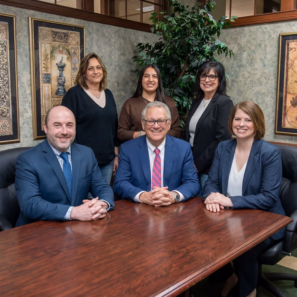 Six business professionals smiling around a wooden conference table in an office setting.