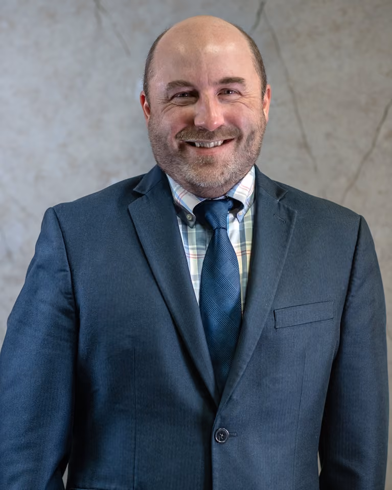 Smiling bald man with beard wearing blue suit jacket, plaid shirt, and blue tie against a gray background.