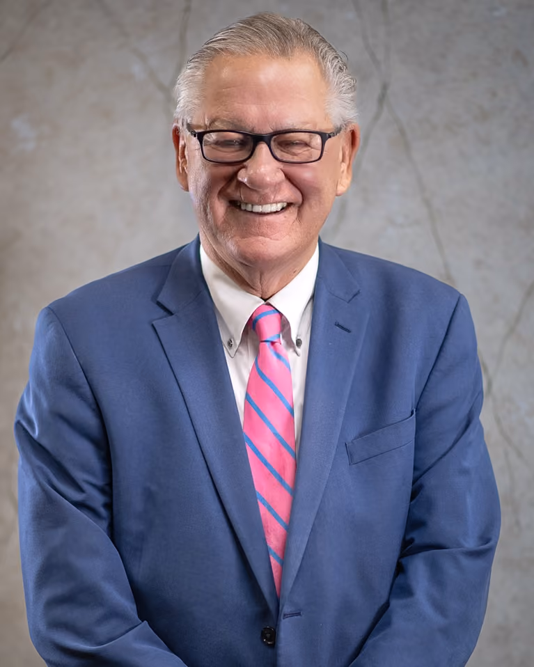 Smiling older man with glasses wearing a blue suit jacket and pink striped tie against gray background.