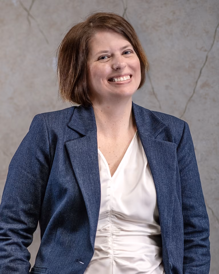 Smiling woman with short brown hair wearing a blue blazer and white blouse against a neutral background.