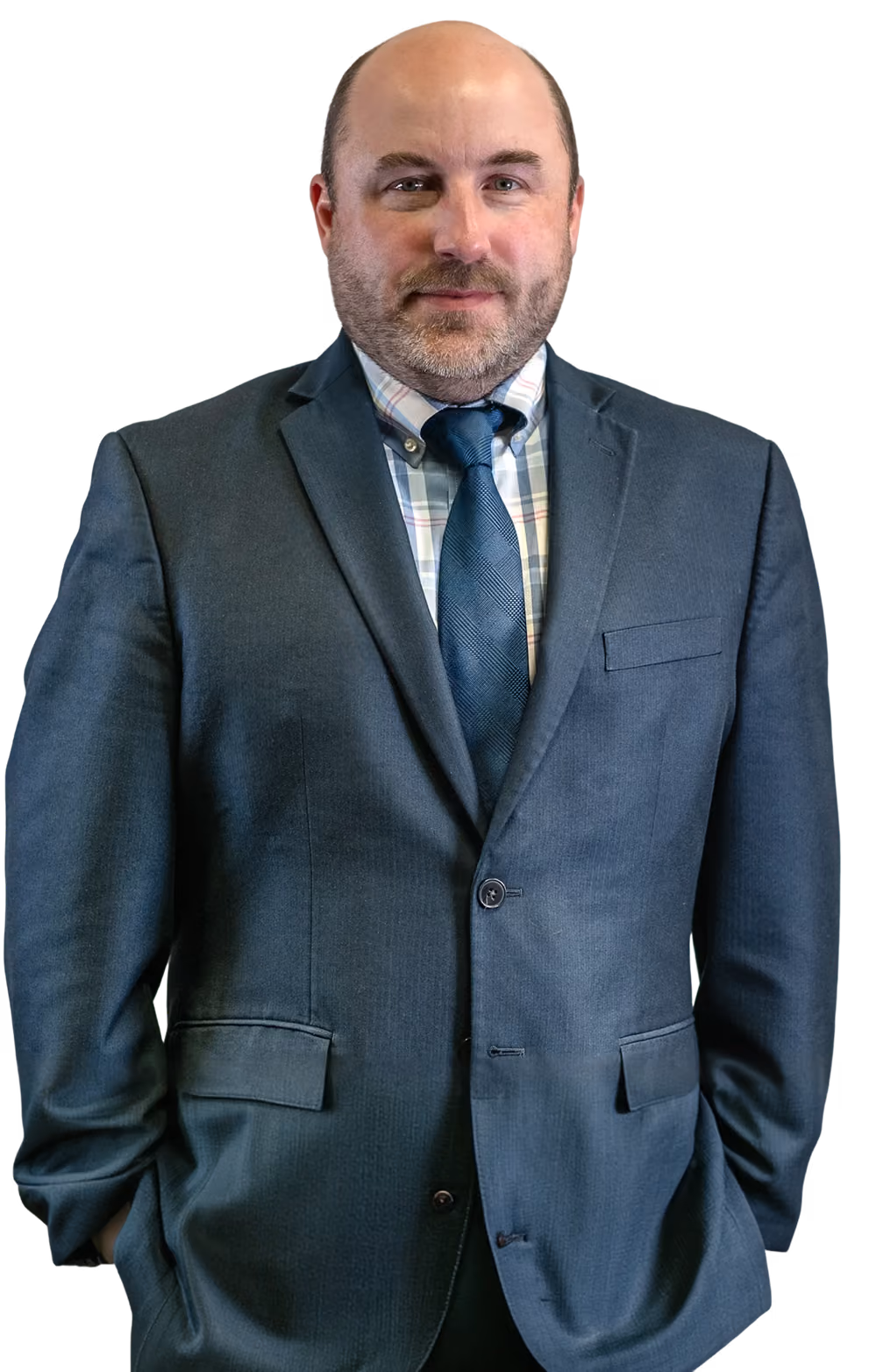 Man in a dark suit, plaid shirt, and blue tie standing with hands in pockets.