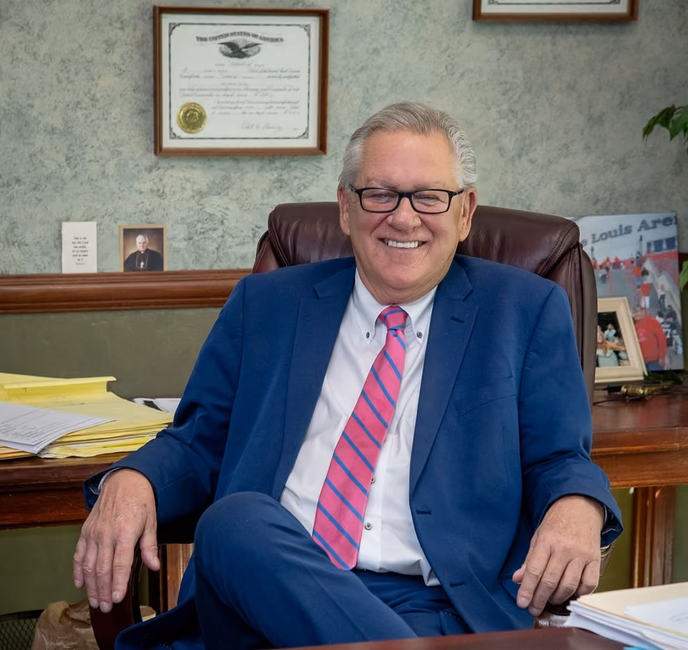 Smiling older man in a blue suit and pink striped tie sitting in an office chair at a desk.