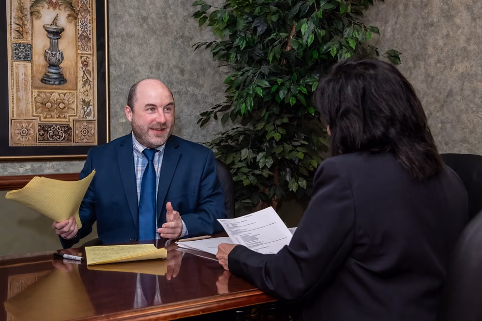 Man in suit holding yellow papers talking to a woman across a desk in an office setting.