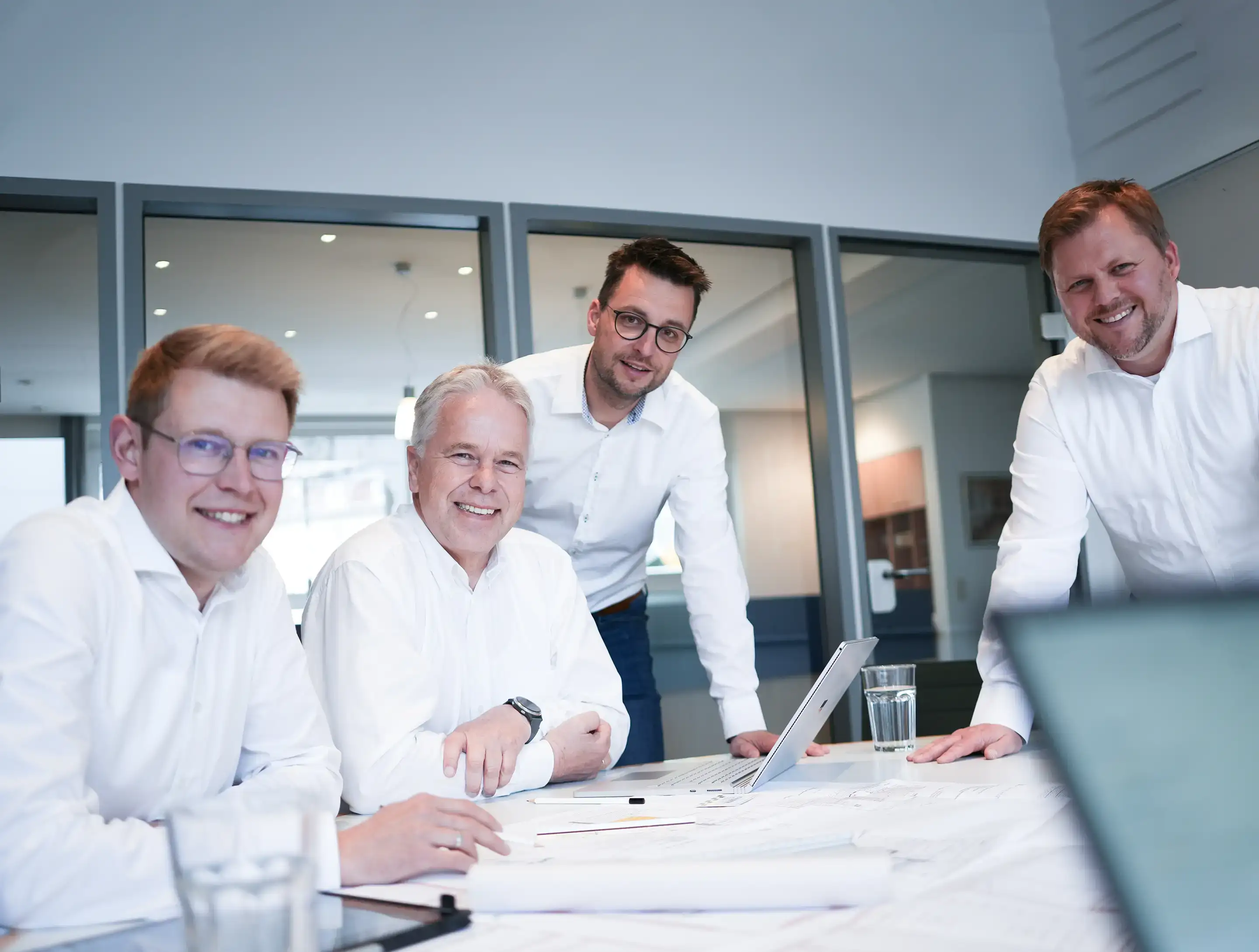Vier Geschäftsleute in weißen Hemden arbeiten gemeinsam an einem Tisch mit Laptop und Plänen.