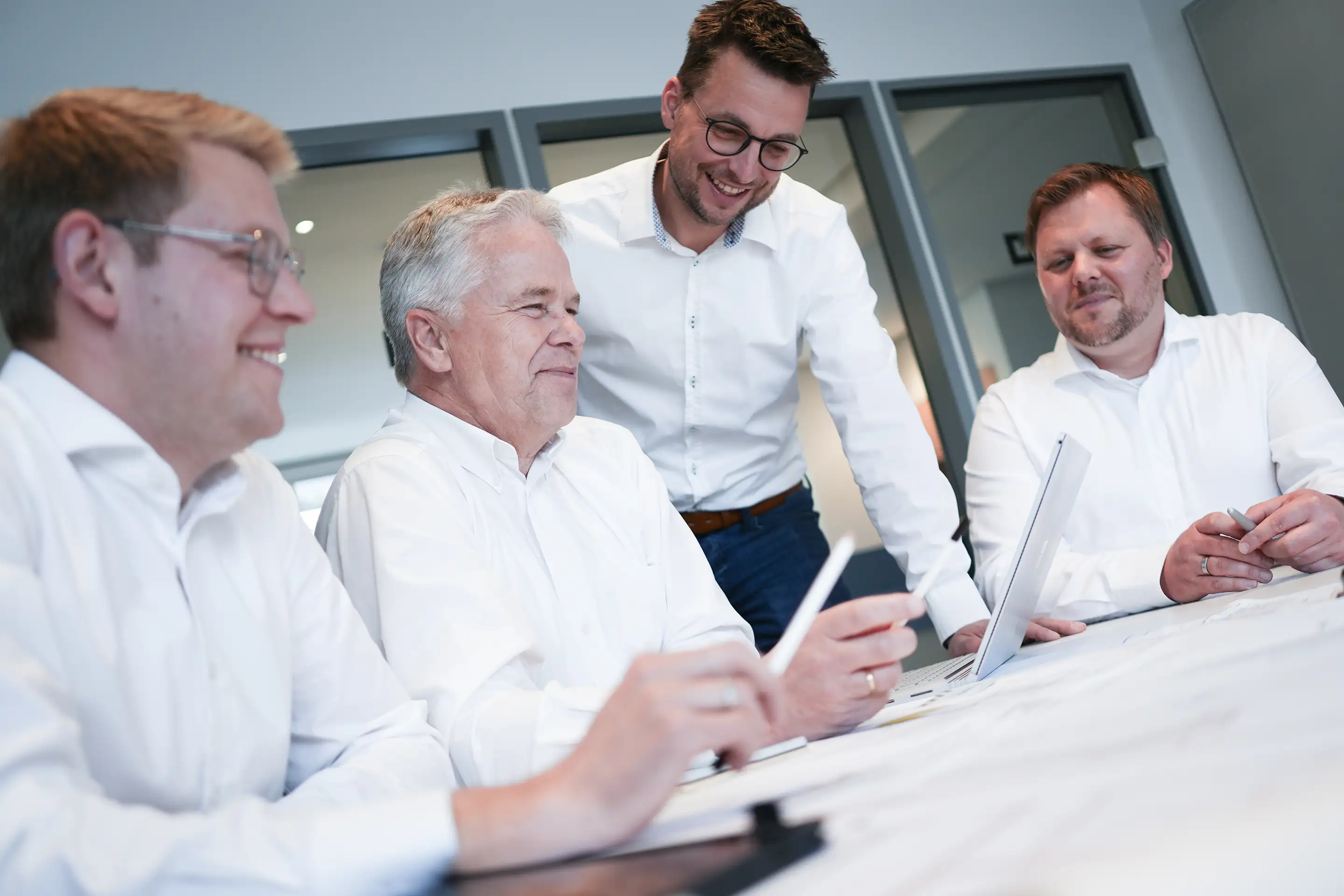 Vier Männer in weißen Hemden sitzen zusammen und diskutieren über Dokumente am Tisch.
