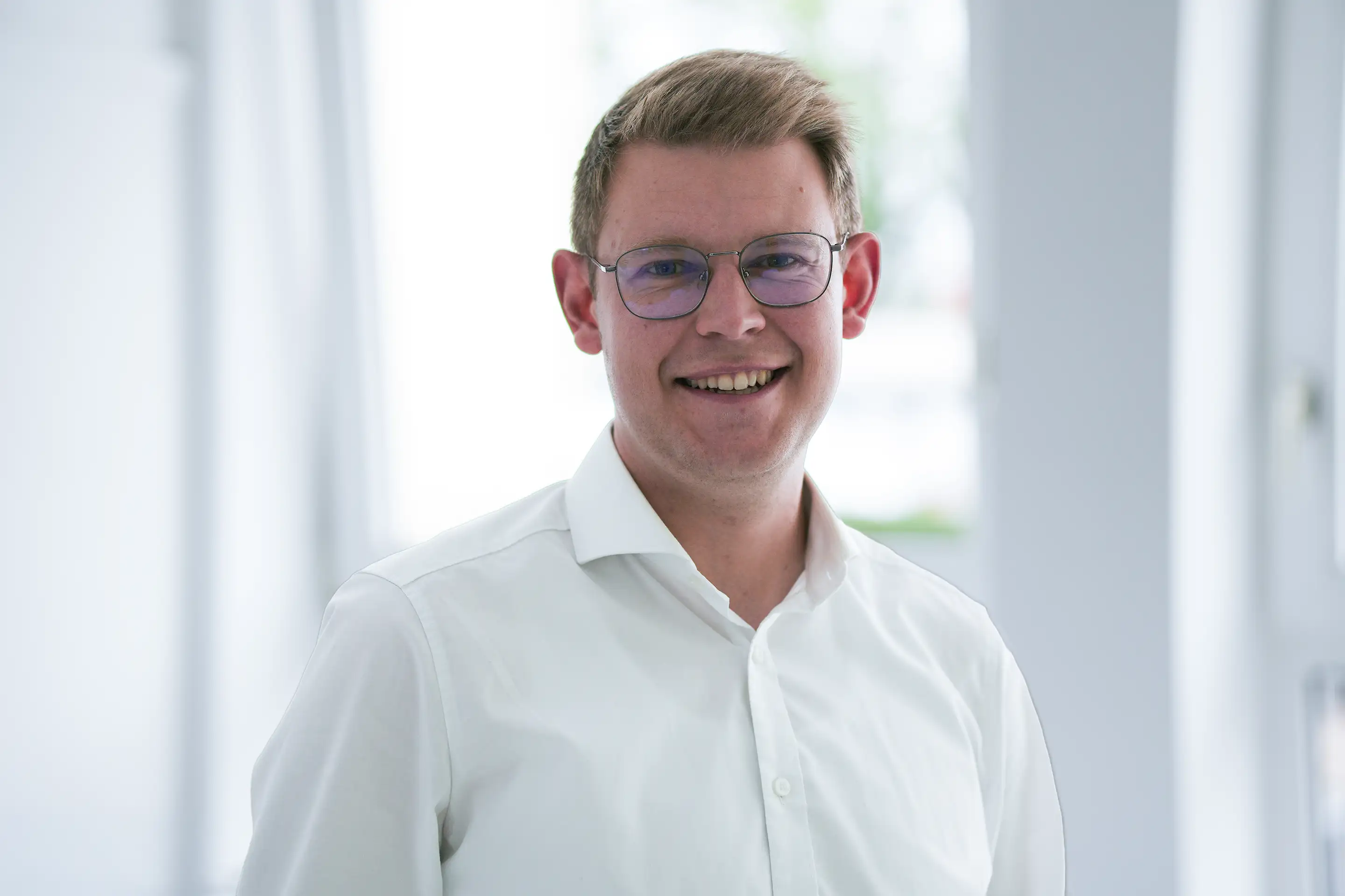 Lächelnder Mann mit Brille und weißem Hemd vor hellem unscharfem Hintergrund.