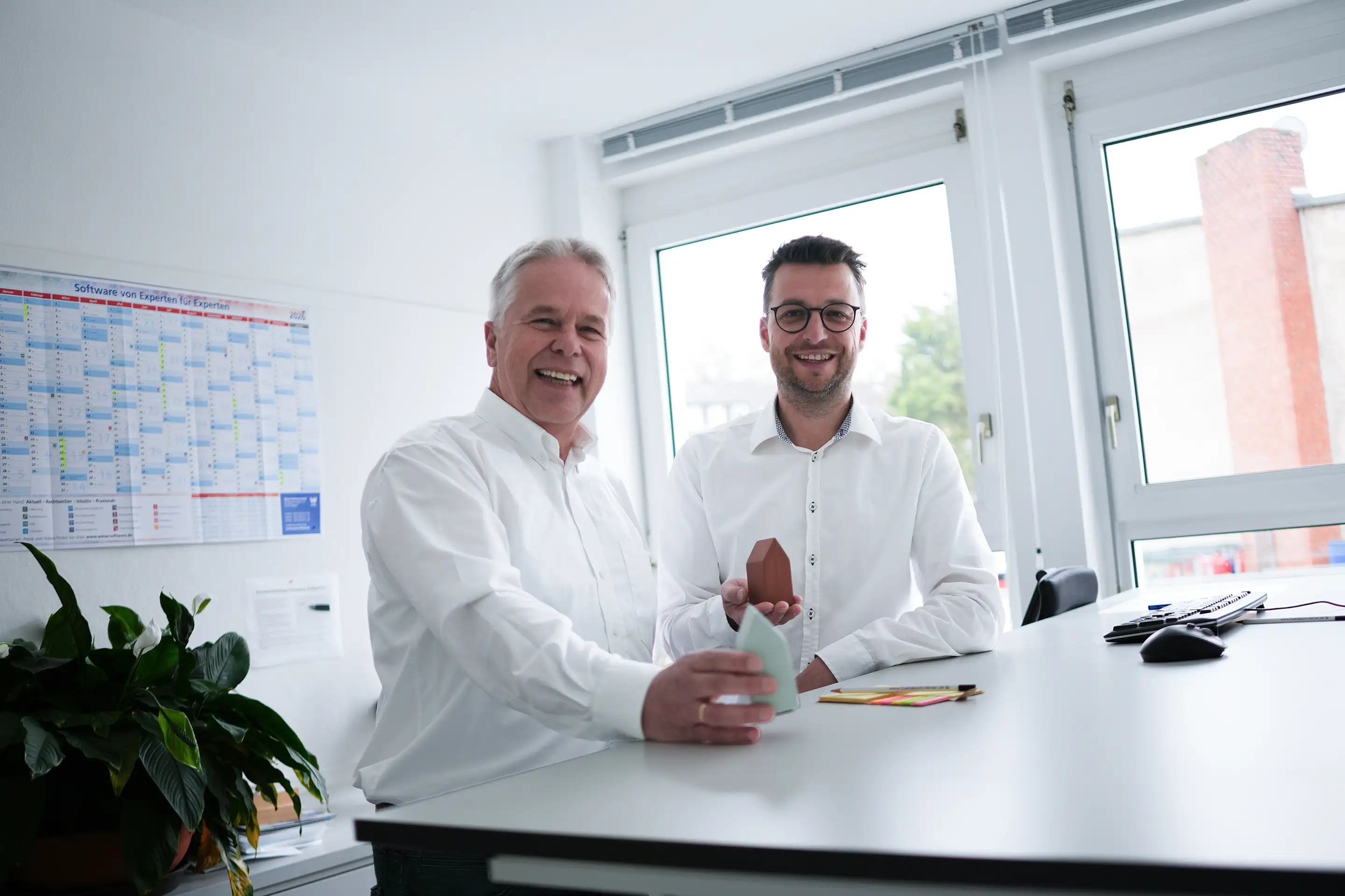 Zwei Männer in weißen Hemden lächeln und halten kleine Hausmodelle am Schreibtisch in einem Büro.