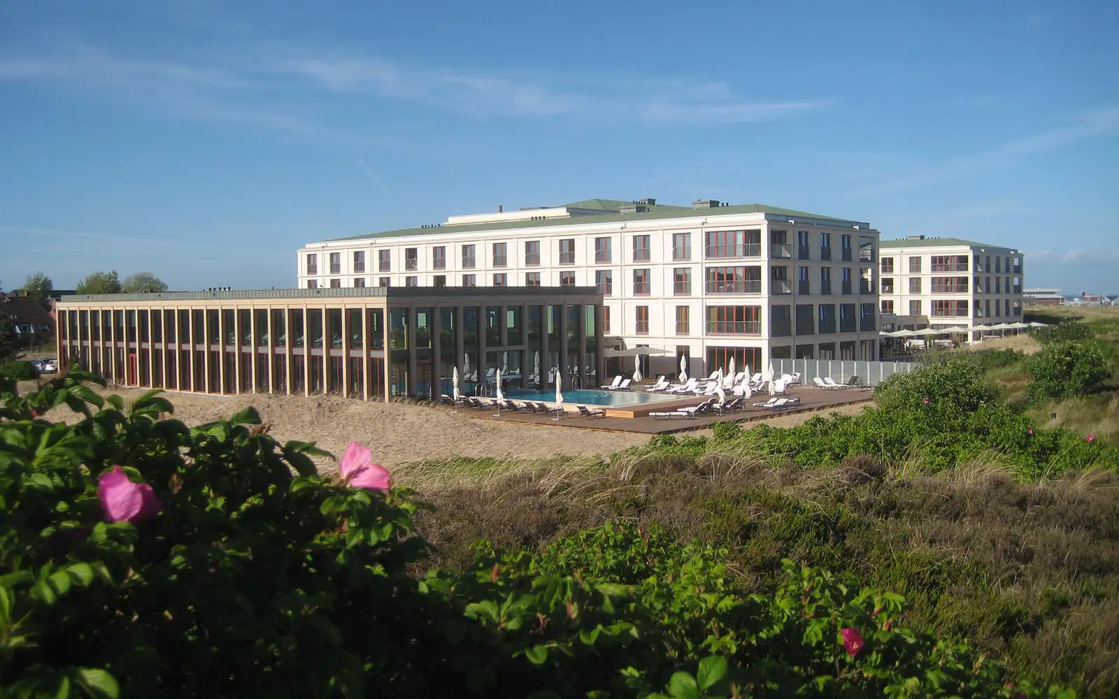 Modern hotel by sandy dunes with outdoor pool, sun loungers, and blue sky in the background.