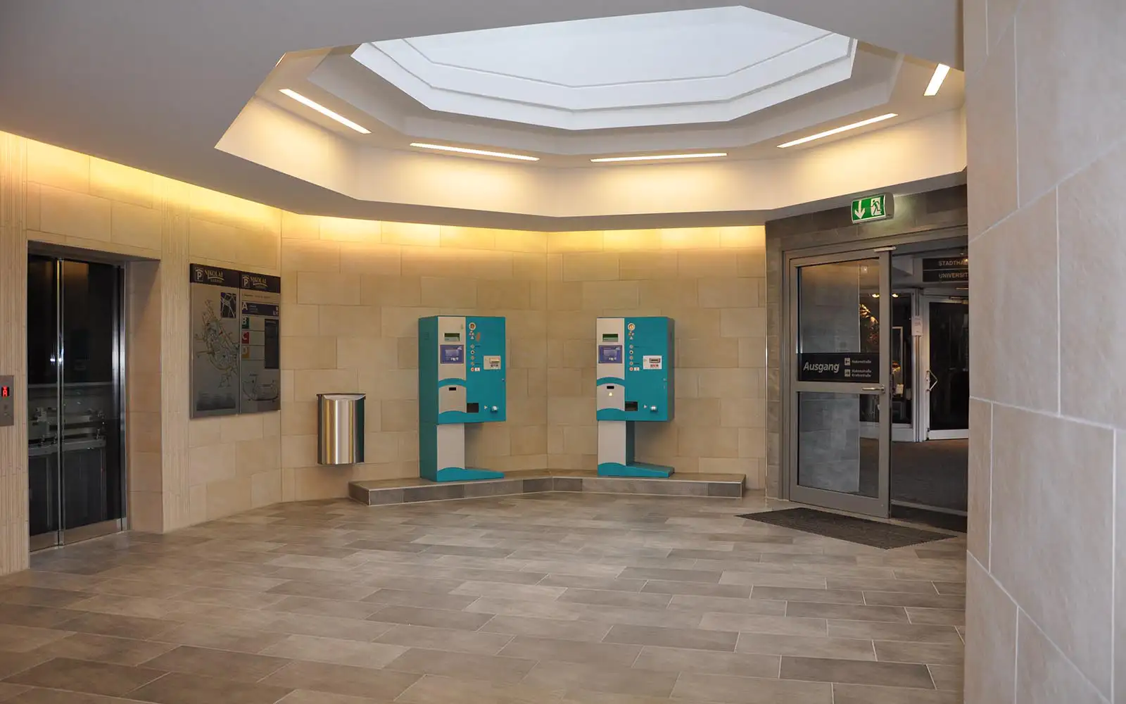 Helle Halle mit zwei blauen Automaten, Müllbehälter, Fahrstuhl und Ausgangstür mit Notausgangsschild.