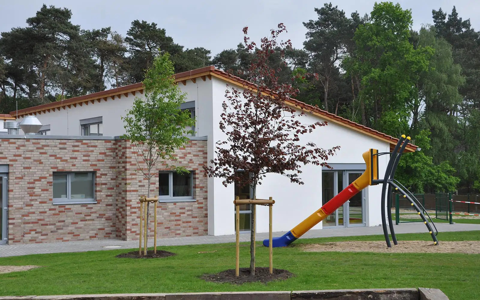Gebäude mit gemauertem Teil und weißer Fassade, davor bunte Rutsche und zwei junge Bäume.