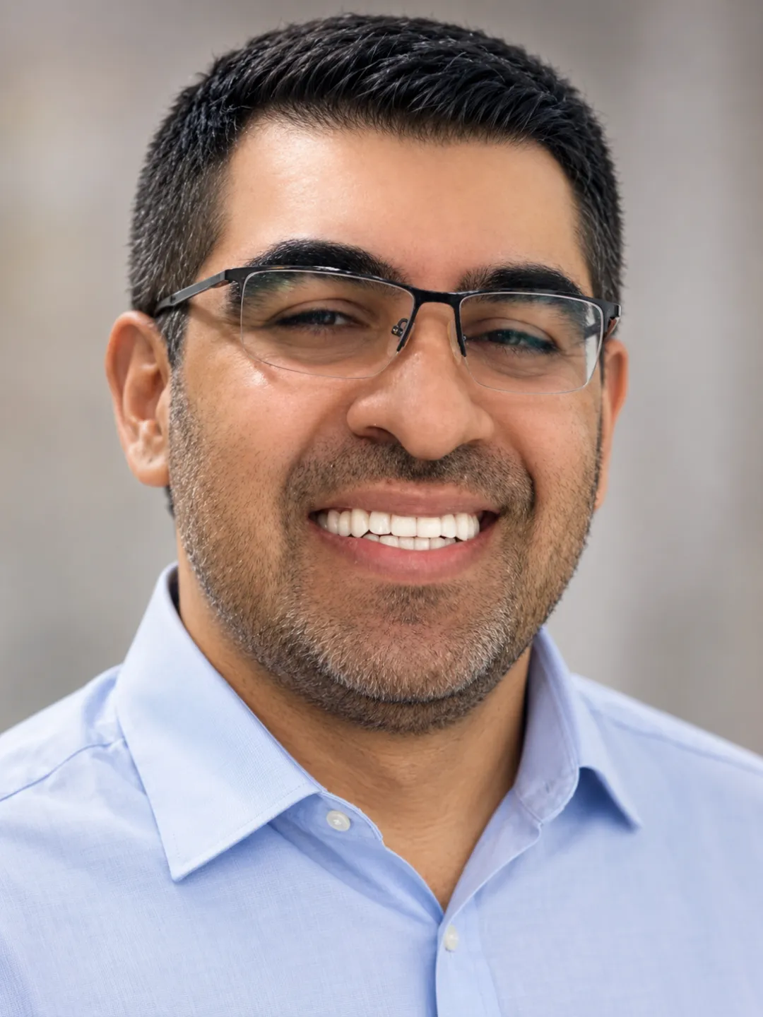 Smiling man with glasses and a light blue collared shirt against a blurred background.