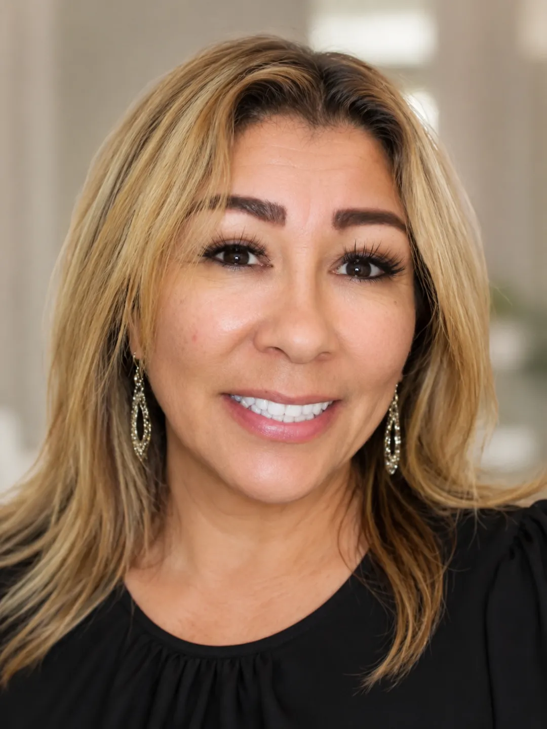 Smiling woman with long blonde hair wearing gold earrings and a black top against blurred background.