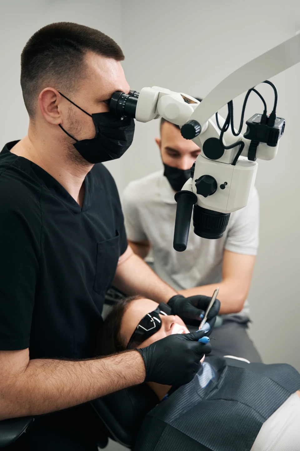 Dentist wearing black mask using dental microscope to treat patient with assistant nearby.