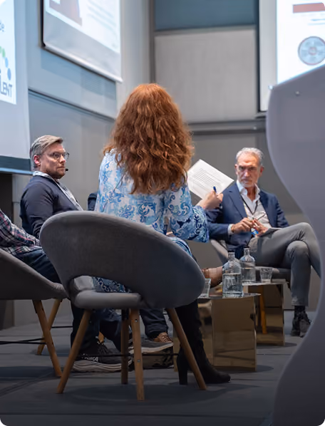 Tre personer i paneldiskusjon på konferanse, med en kvinne i midten sett bakfra.