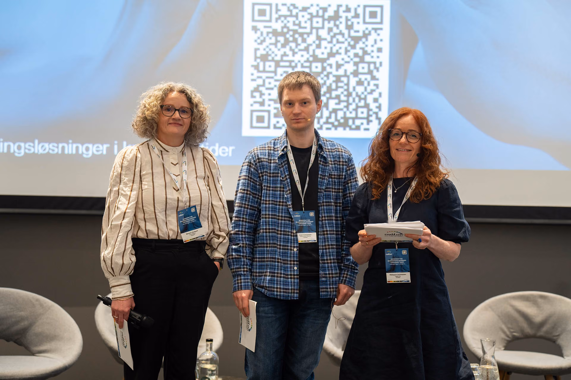 Tre personer står foran en skjerm med QR-kode; de holder papirer og har konferansemerker rundt halsen.