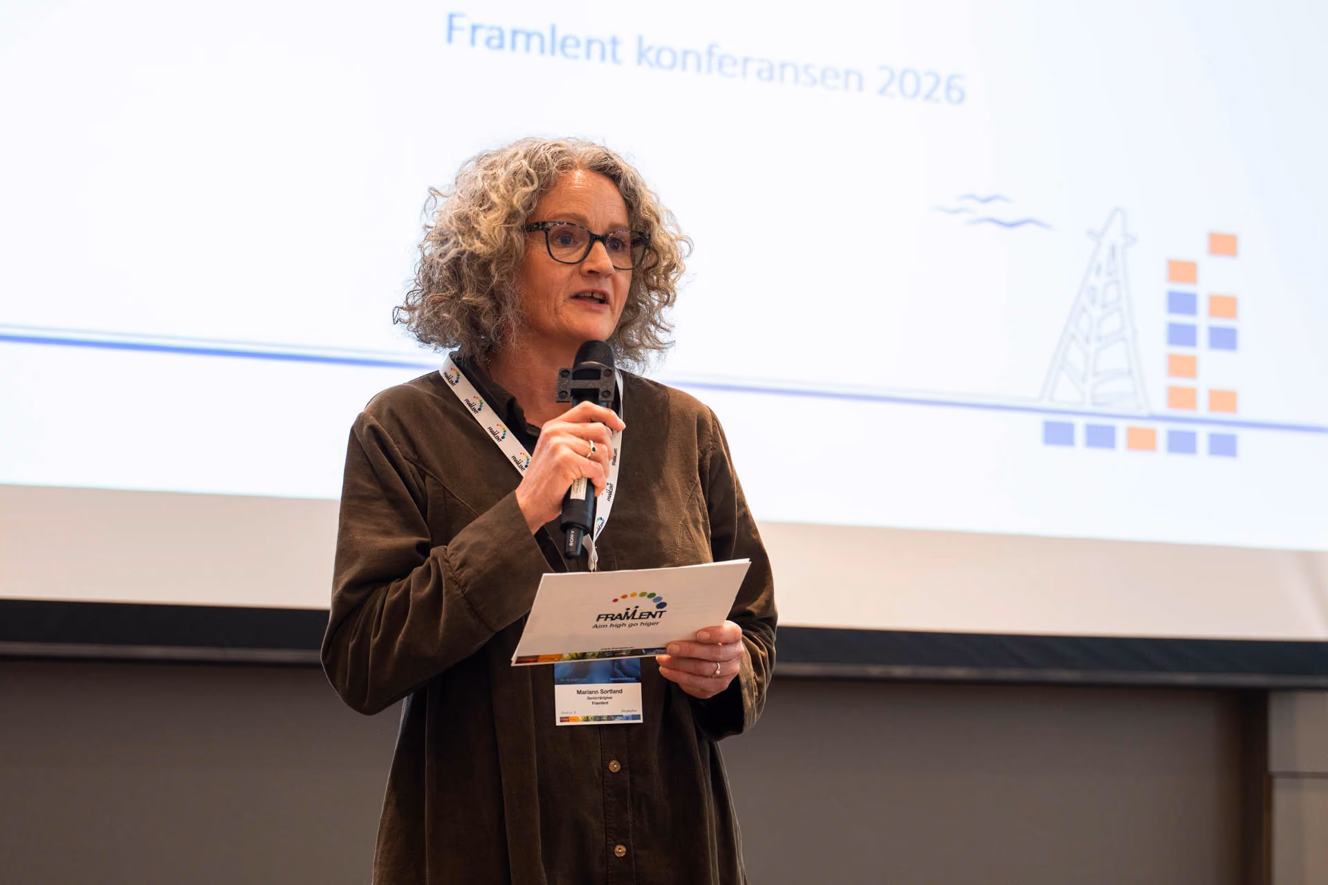 Kvinne med briller holder mikrofon og papir foran skjerm som viser FramIent konferansen 2026.