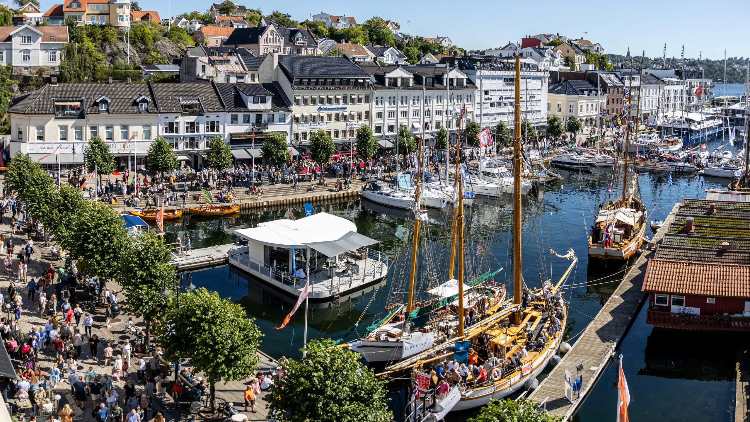 Folkefylt havneområde med seilbåter og bygninger i bakgrunnen på en solrik dag.