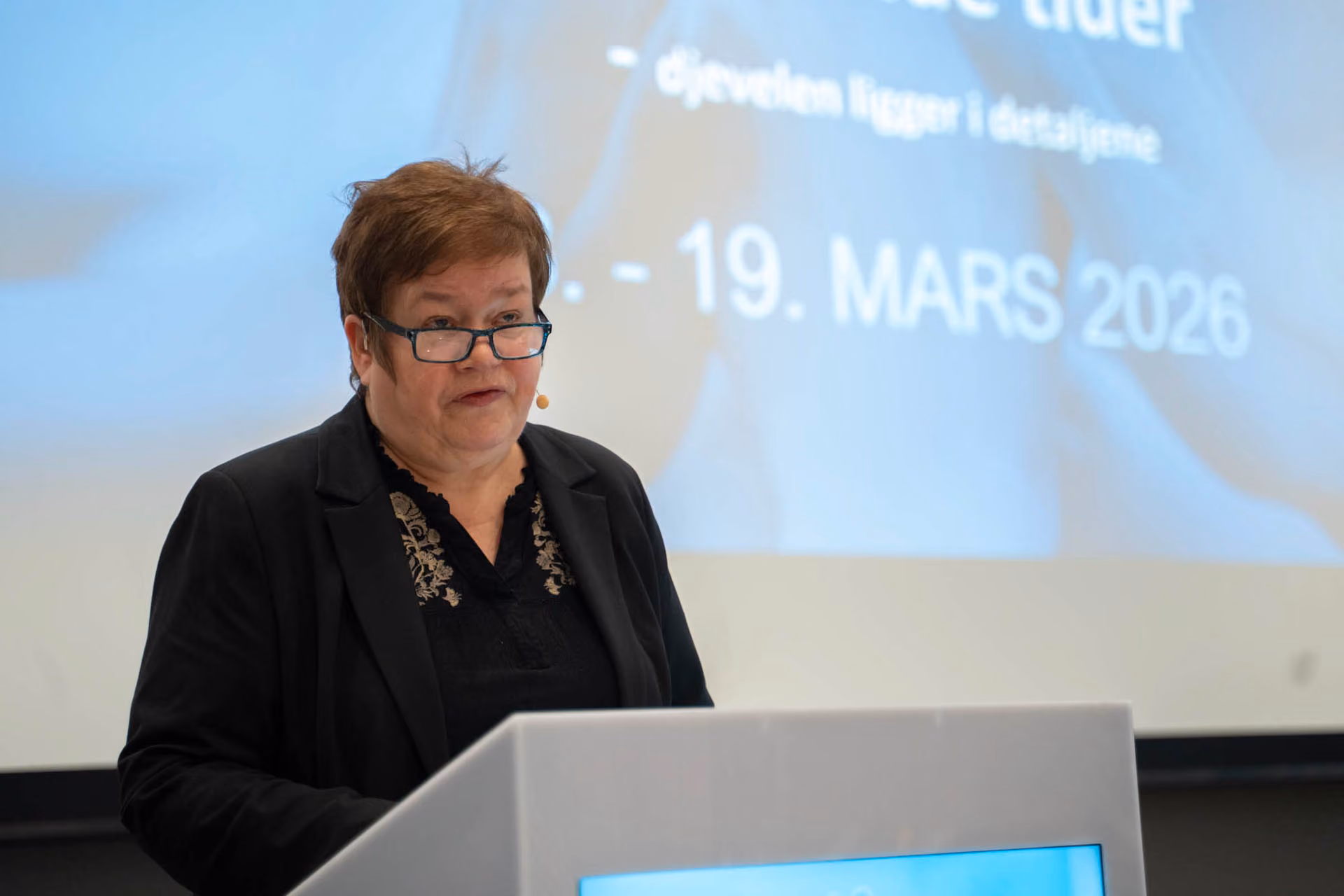 Kvinne med briller som holder tale foran talerstol med skjerm og blå bakgrunn med tekst.