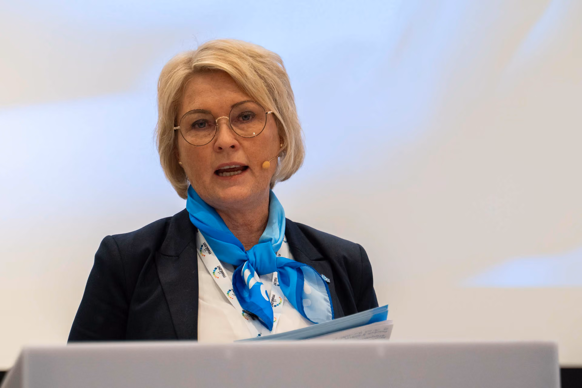 Kvinne med kort blondt hår og briller holder talepapirer og snakker foran mikrofon.