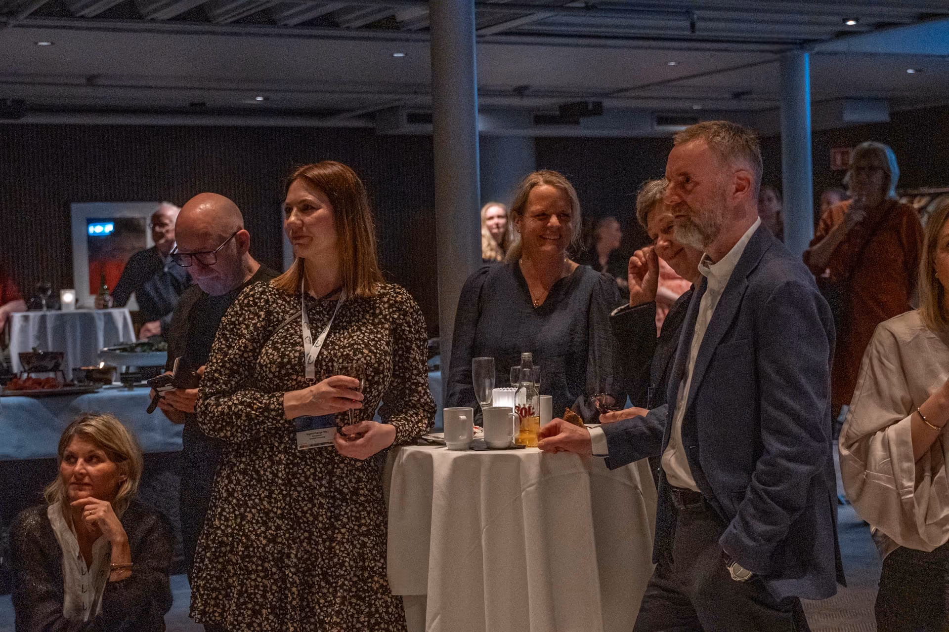 Folk i en gruppe står rundt et høyt bord med drikke og ser ut mot noe utenfor bildet.