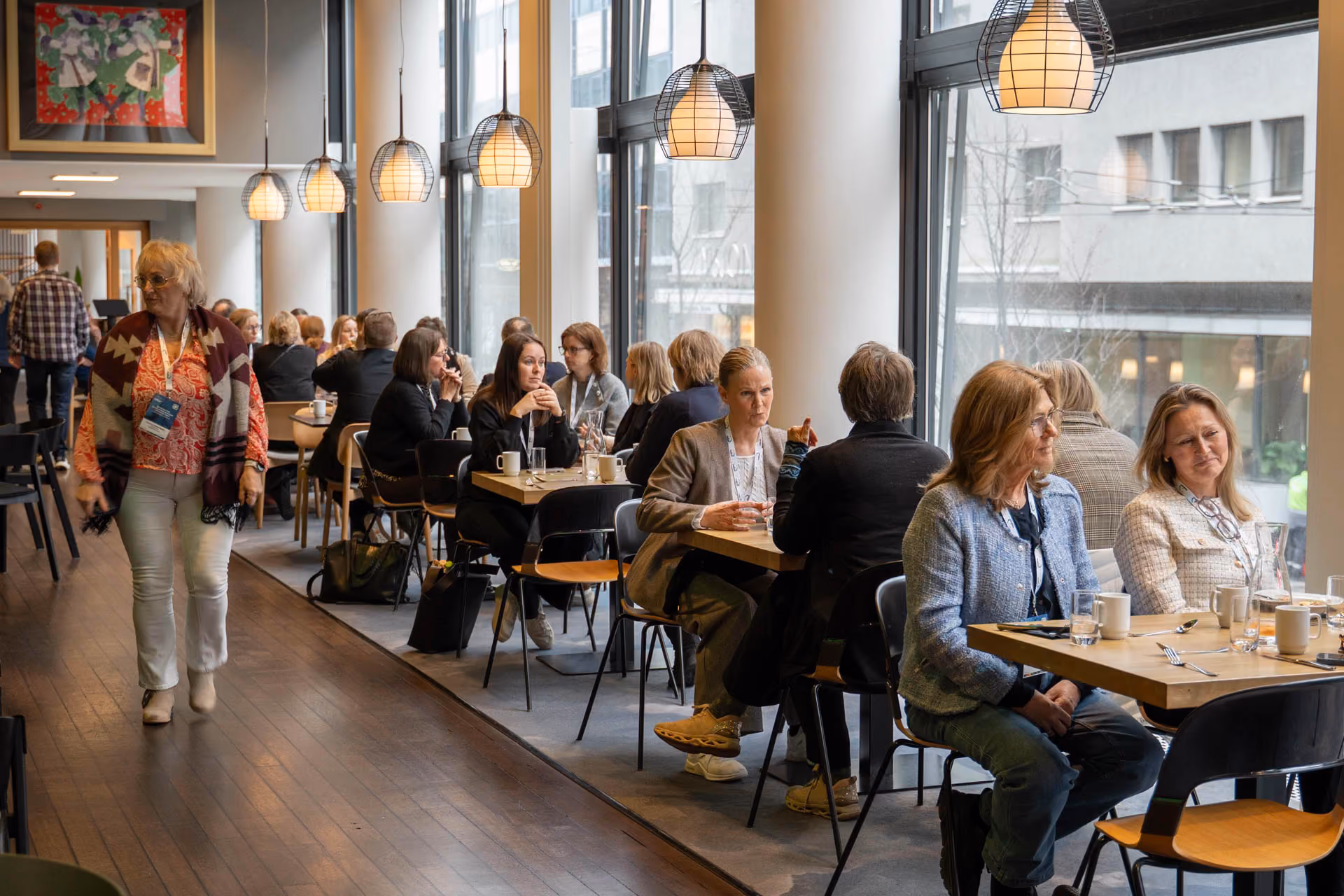 Folk sitter og snakker rundt bord i et lyst kaféområde med store vinduer og hengende lamper.