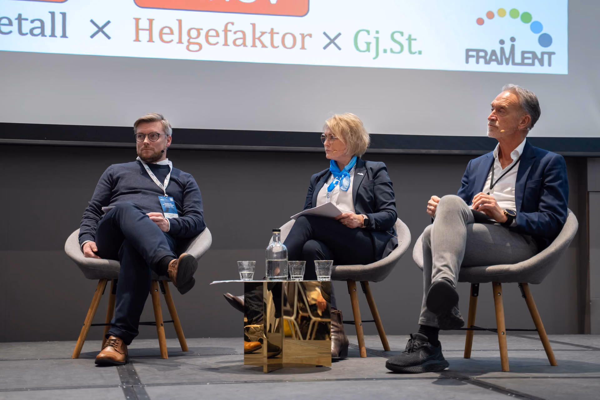 Tre personer i en paneldiskusjon på scenen med et bord med vannflaske og glass foran dem.