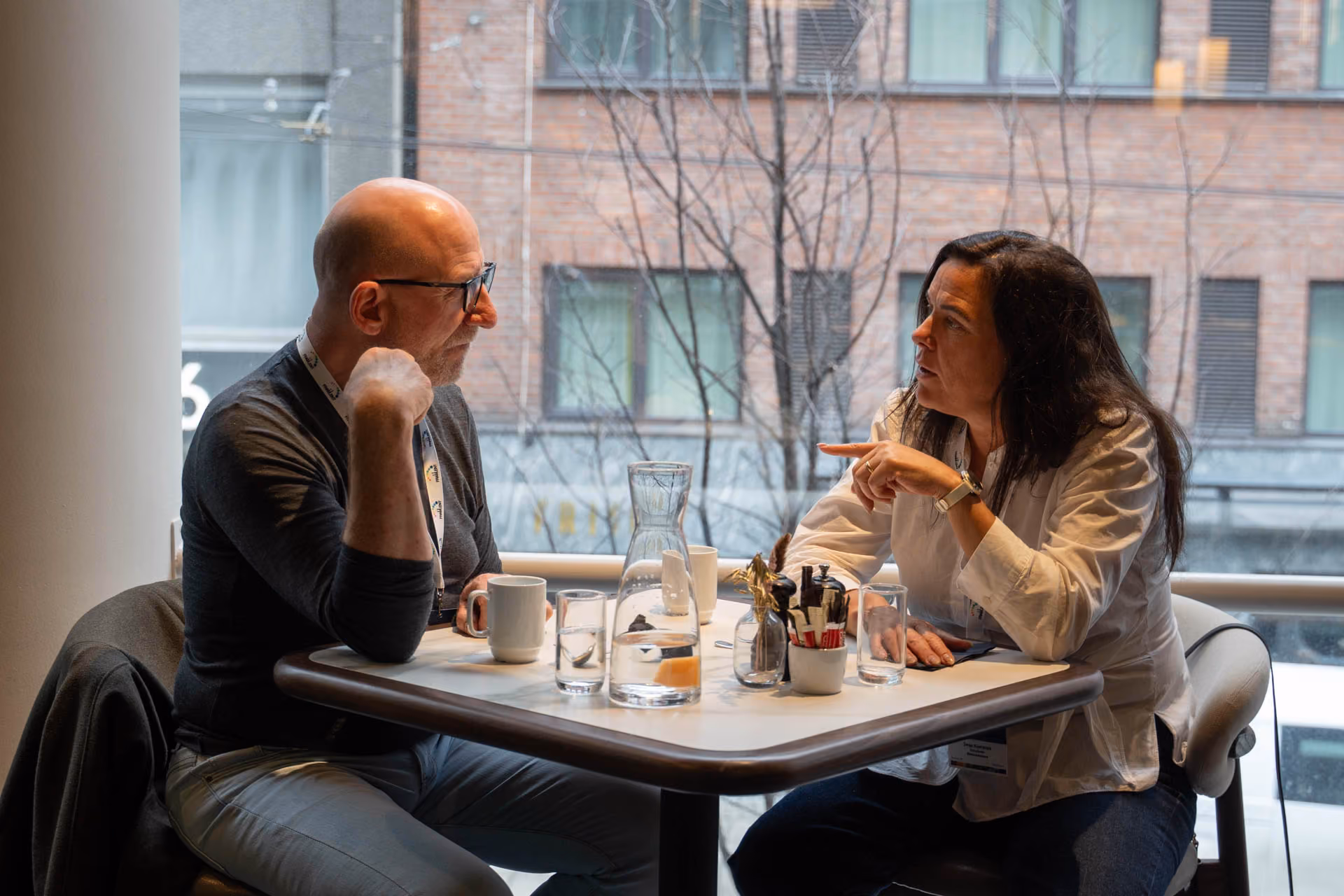 To voksne mennesker sitter ved et bord og har en samtale med glass og kaffekopper foran seg.