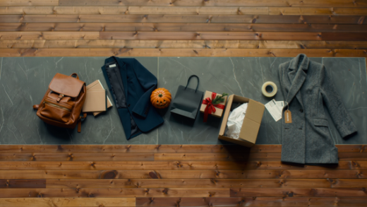 Wooden table with a checked blazer, a leather backpack, folded knitted jumpers, a scarf, a pumpkin, a wrapped gift and a coat with a winter sale tag, with a clothing rail in the background