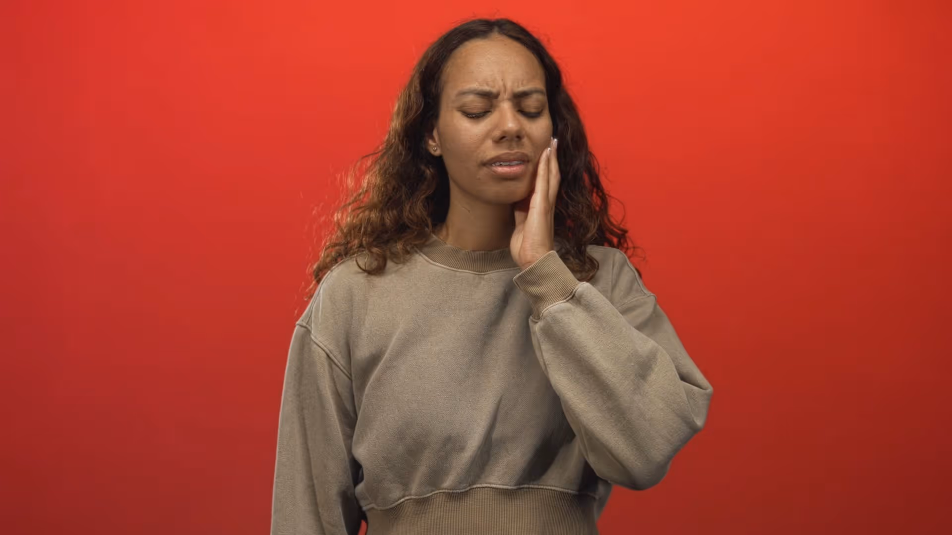 Woman with curly hair in beige sweater holding her cheek with a pained expression on red background.
