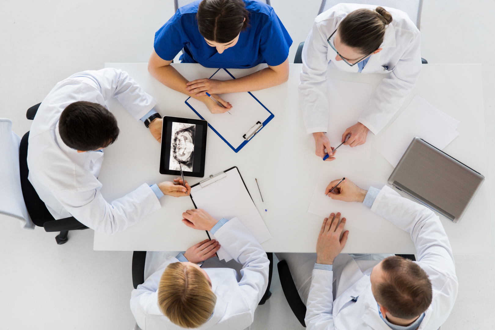 Five medical professionals in white coats and scrubs reviewing a dental X-ray on a tablet at a table.