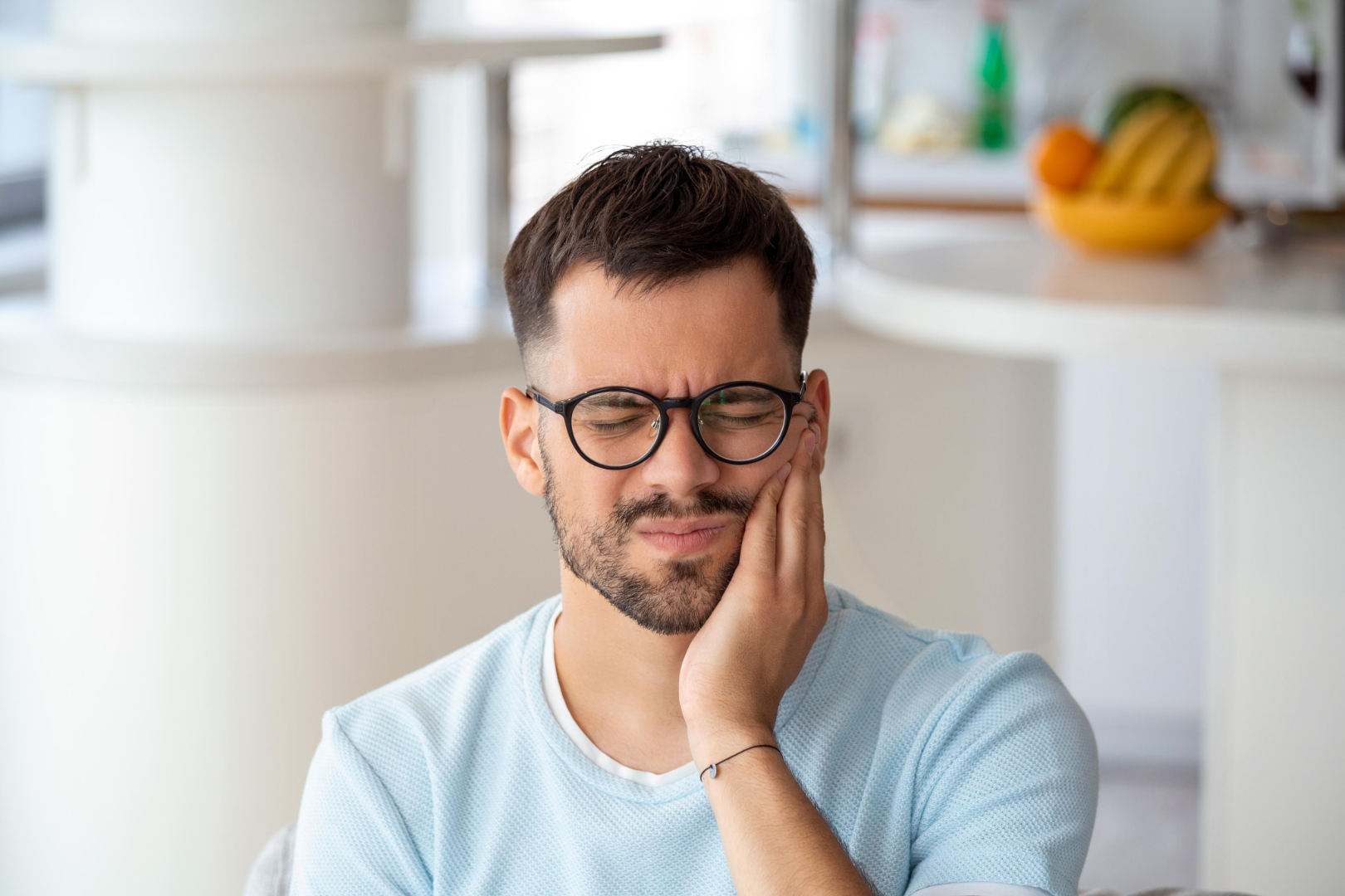 Man with glasses in light blue shirt holding his cheek in pain with eyes closed indoors.